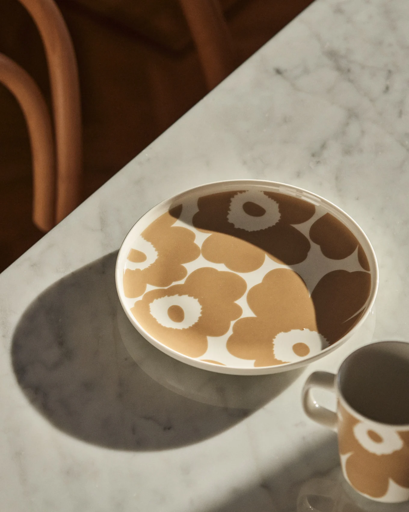 A plate with a tan floral pattern and a matching mug on a marble table with sunlight and shadows.