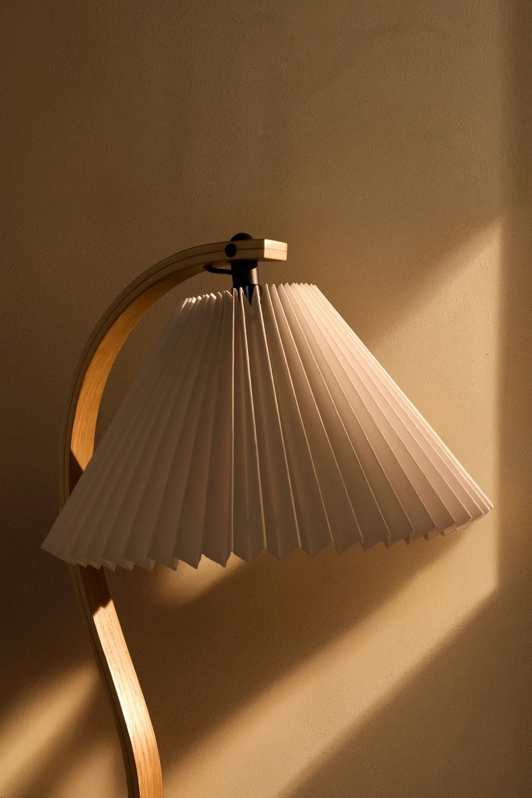 A lamp with a white pleated shade and a curved wooden base, casting warm light on a textured wall.