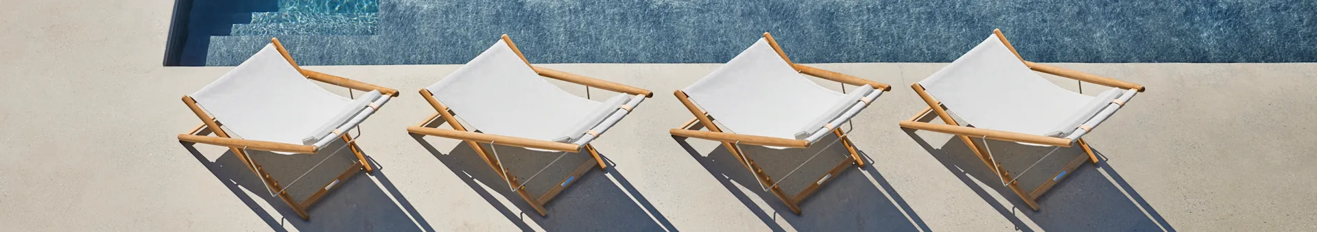 Four light-colored sling lounge chairs with wooden frames by a blue swimming pool on a sunny day.