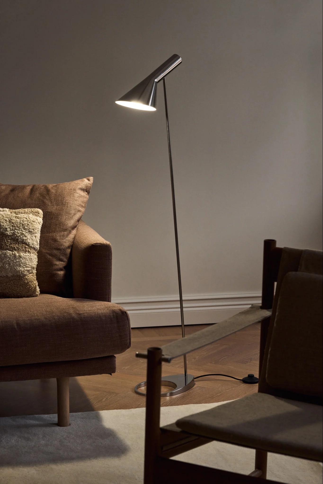 A modern, sleek metallic floor lamp with a conical shade illuminating a corner with a brown sofa and a wooden chair.