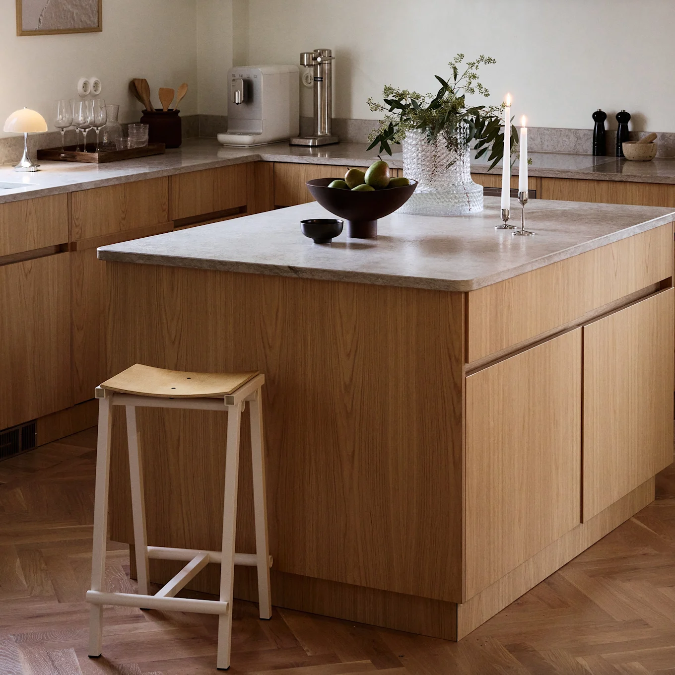 Modern kitchen with light wood cabinets, an island, and a white stool. Green fruit and lit candles adorn the island.