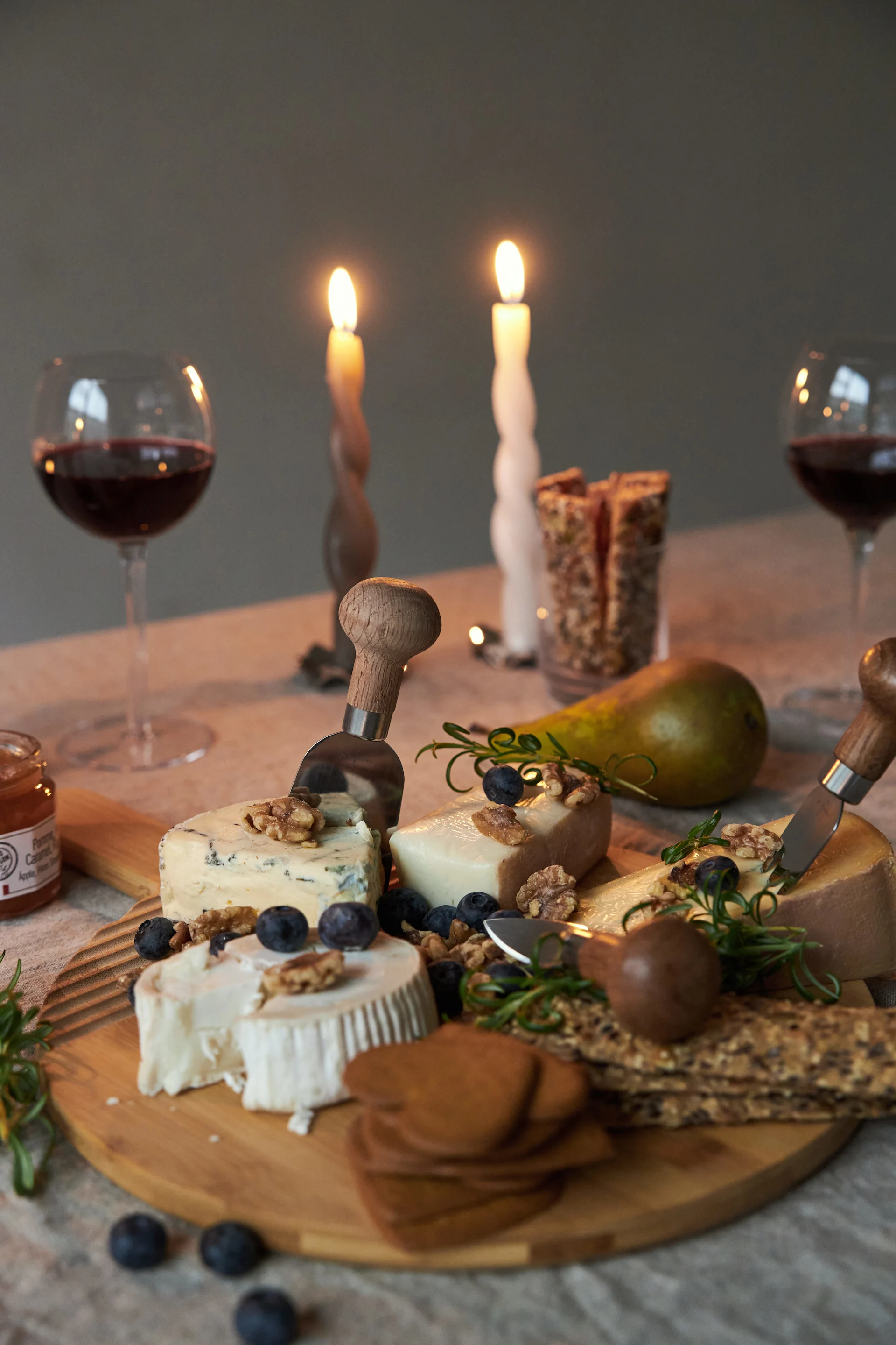 A cheese board with various cheeses, blueberries, walnuts, crackers, and jam, served with red wine and lit candles.