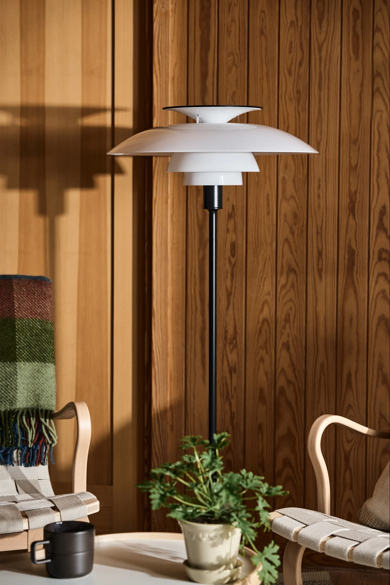 White layered floor lamp next to two wooden chairs and a plant against a wood-paneled wall.