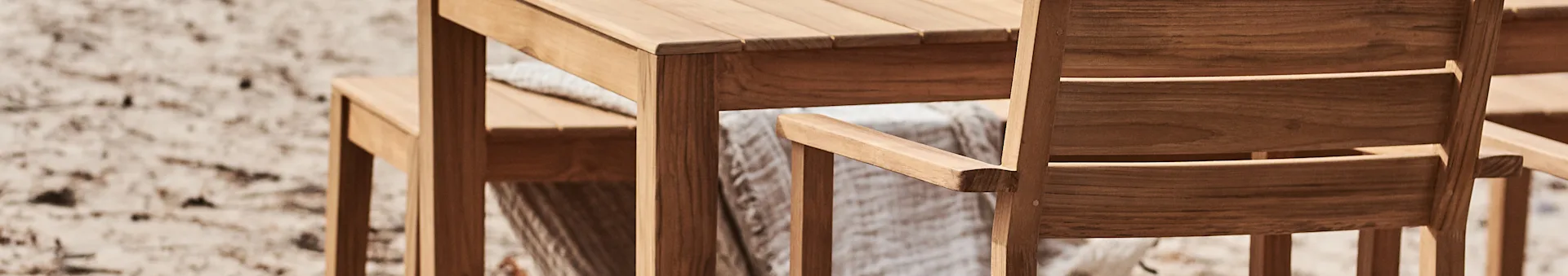 Close-up of a wooden outdoor table and chair set, with a light throw draped over a chair, on a blurred sandy background.