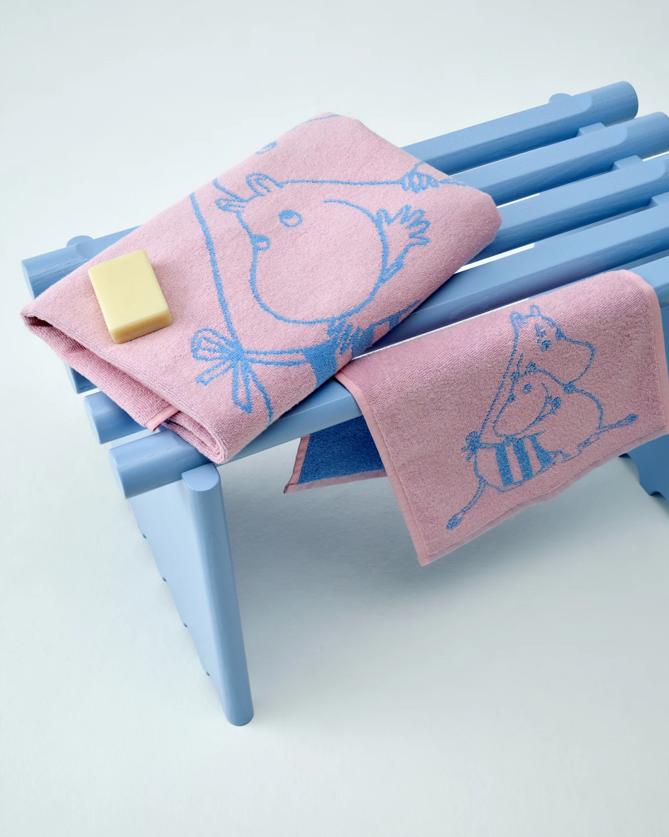 Two pink Moomin towels with blue patterns, one folded with a bar of soap, on a light blue bench.
