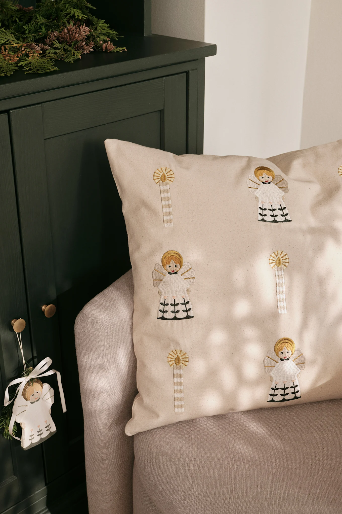 Light beige cushion with embroidered angels and candles on a grey sofa, next to a dark green cabinet with greenery and a hanging angel ornament.