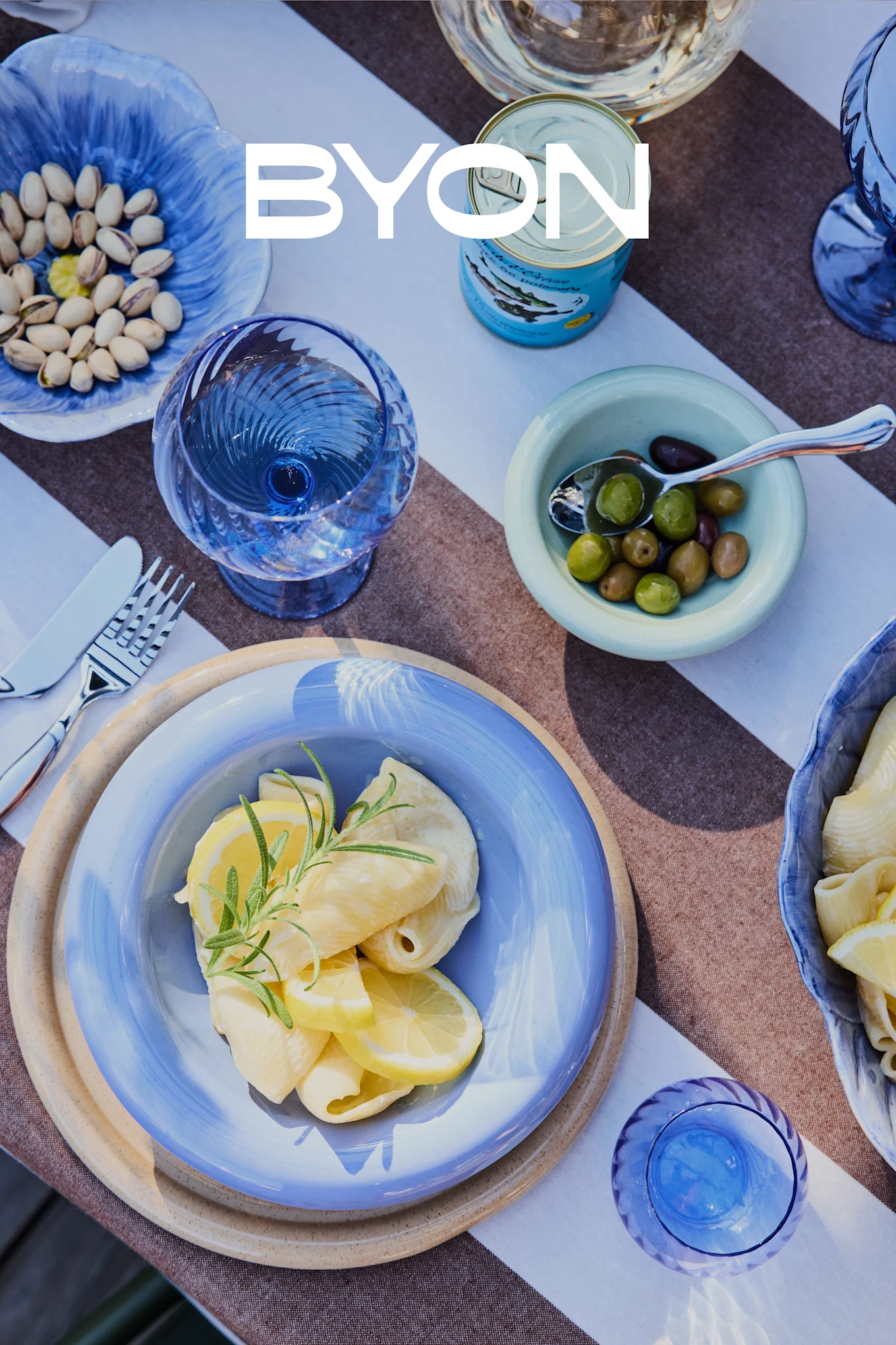 Overhead view of a dining table with blue tableware, including a plate of pasta with lemon and rosemary, bowls of pistachios and olives, and blue glasses. The BYON logo is overlaid.