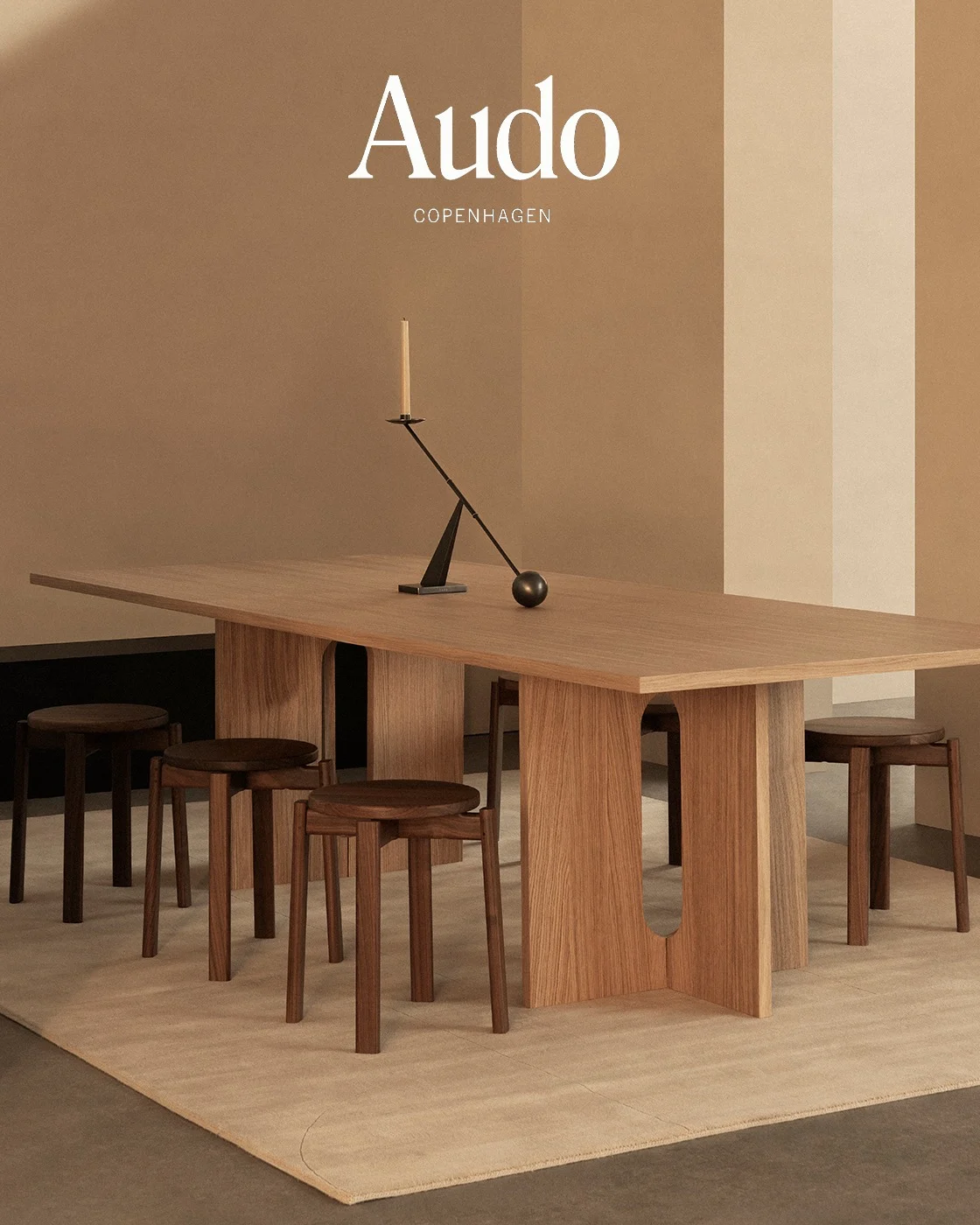 A long light wooden table, dark wooden stools, and a black candlestick with a white candle on a beige rug.