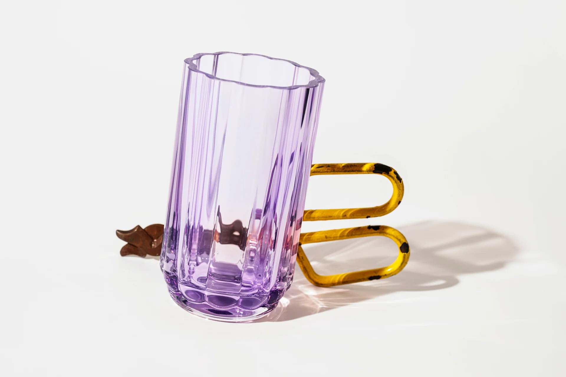 A tall, ribbed purple glass with a wavy rim sits next to a rusty yellow U-shaped handle on a white background.