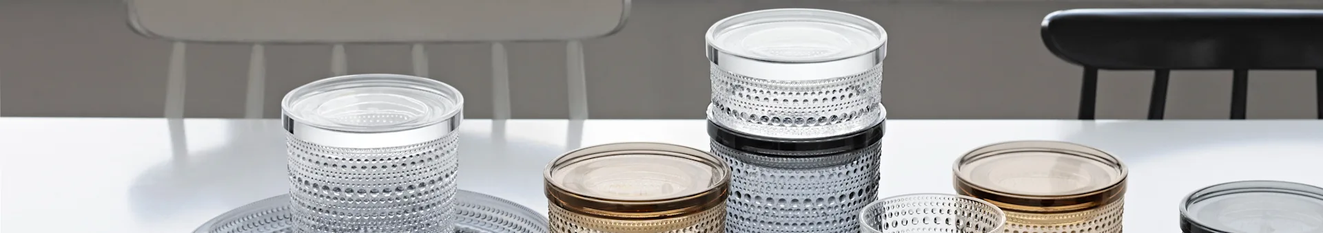 Collection of clear, amber, and dark textured glass containers on a white table with chairs in the background.