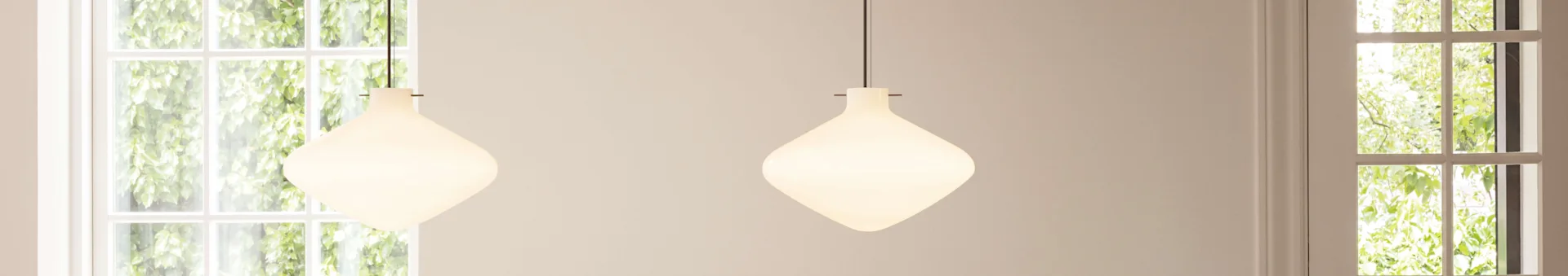 Two unique rounded white glass pendant lights hanging in front of sunlit windows with green foliage.