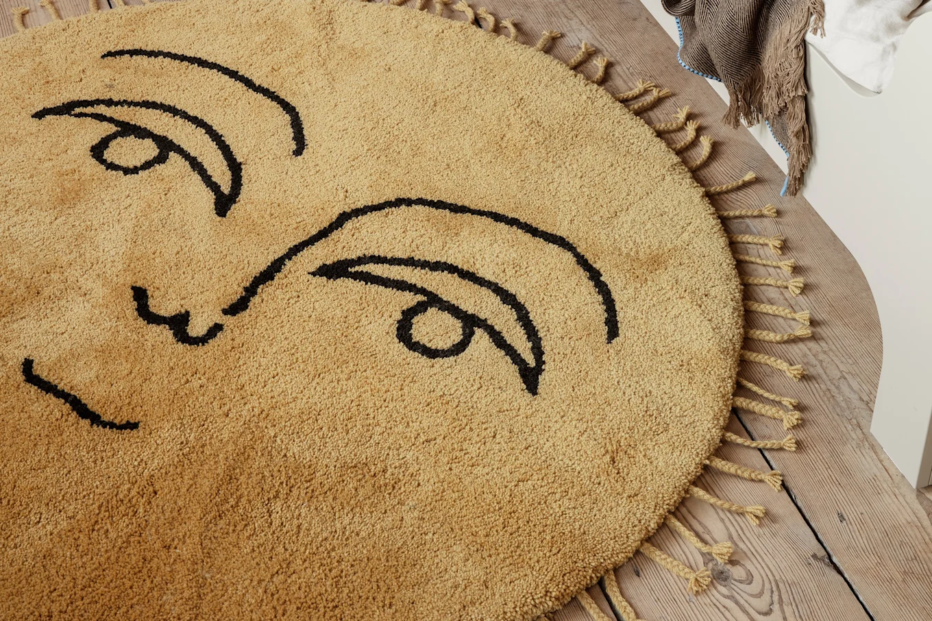 Round tan shaggy rug with black abstract face design and tassels on a wooden floor.
