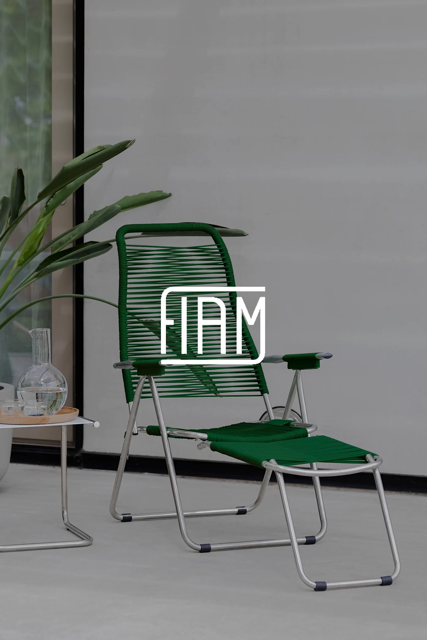 A green reclining chair with a metal frame and footrest, next to a small table with a carafe and glasses. A plant is in the background.