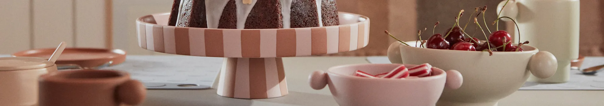 A chocolate cake with white icing on a striped pink and beige cake stand, flanked by bowls of candies and cherries.
