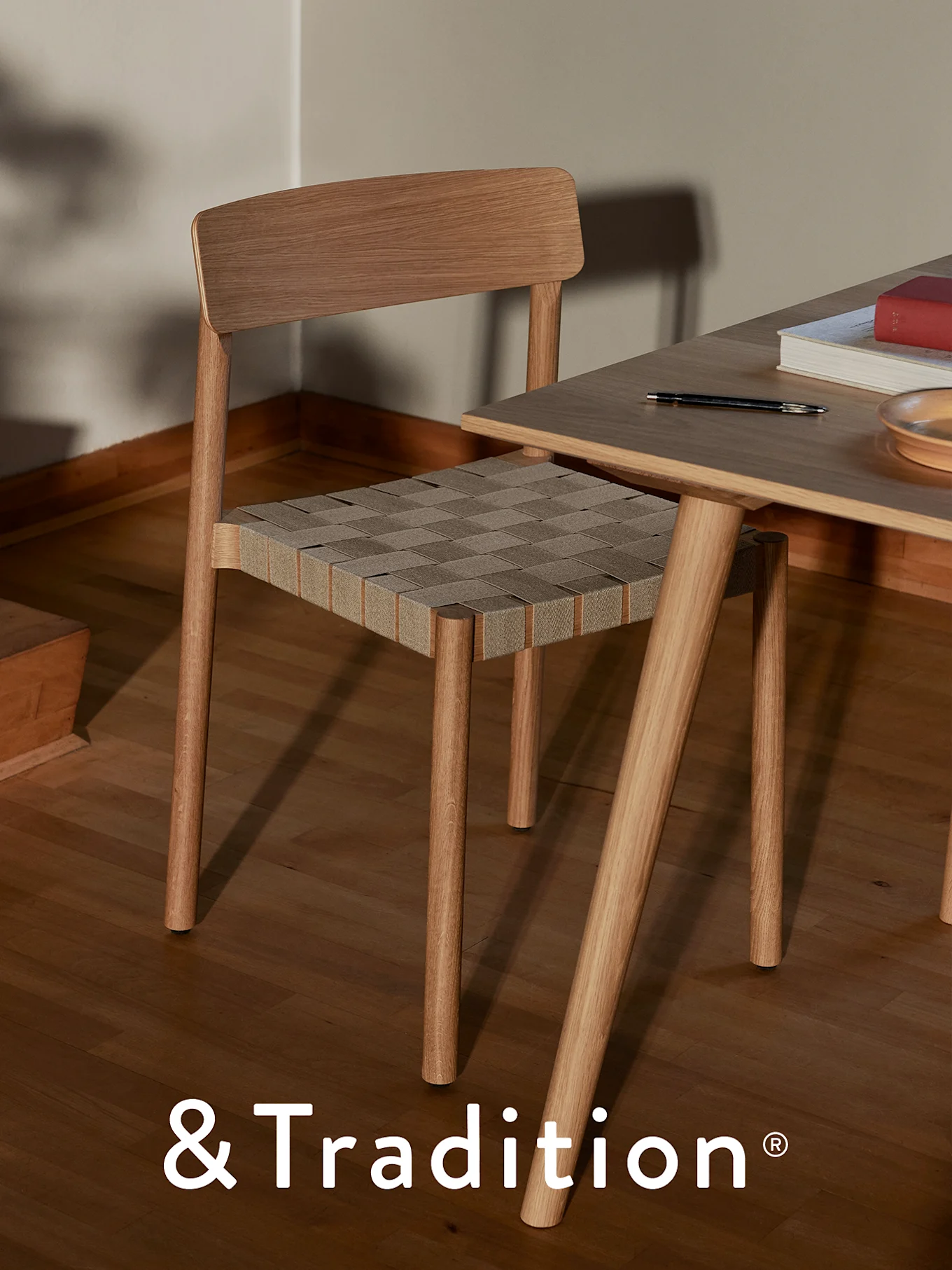 A light wooden chair with a woven tan and gray seat next to a matching wooden table with a pen, book, and plate.