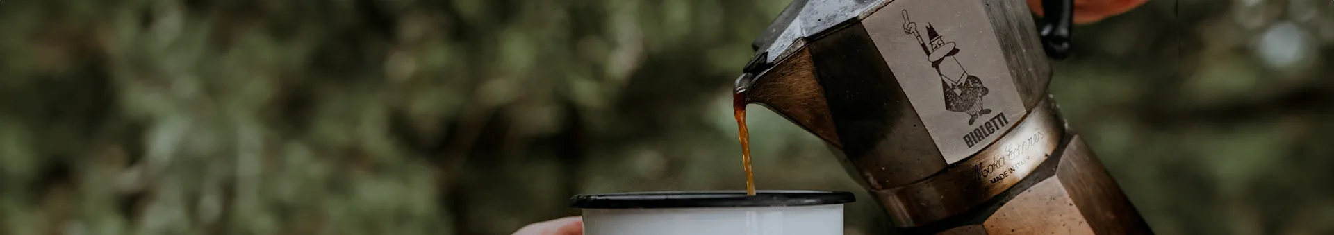 Coffee being poured from a Bialetti Moka Express pot into a white enamel mug.