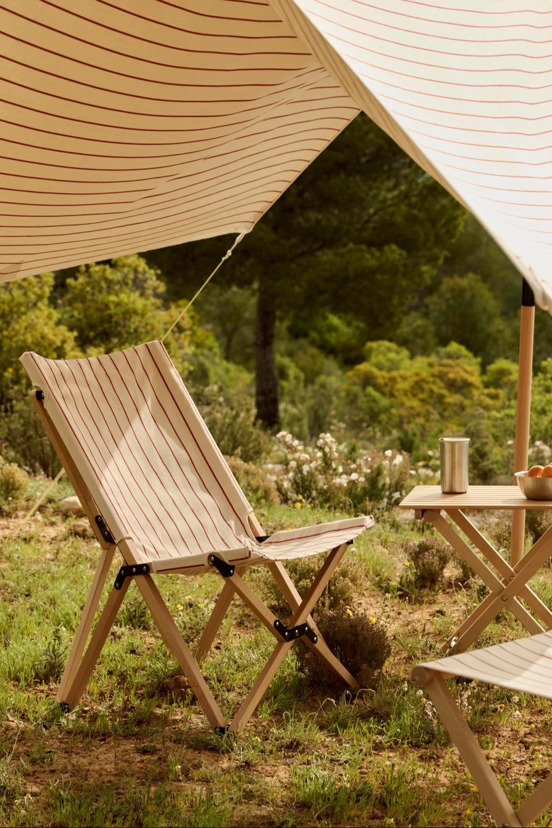A striped canopy shades a striped wooden folding chair and a table with a thermos and oranges in a natural outdoor setting.