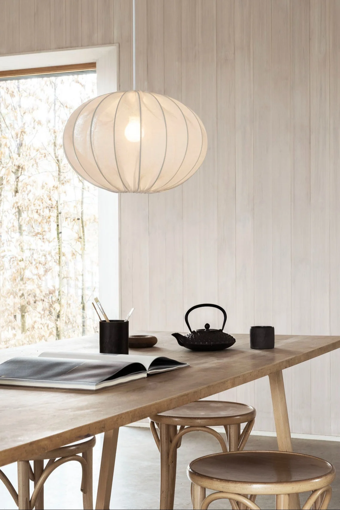 An oval paper lantern pendant light hangs above a wooden table with an open book, a black teapot, and two black mugs. A window overlooks trees.