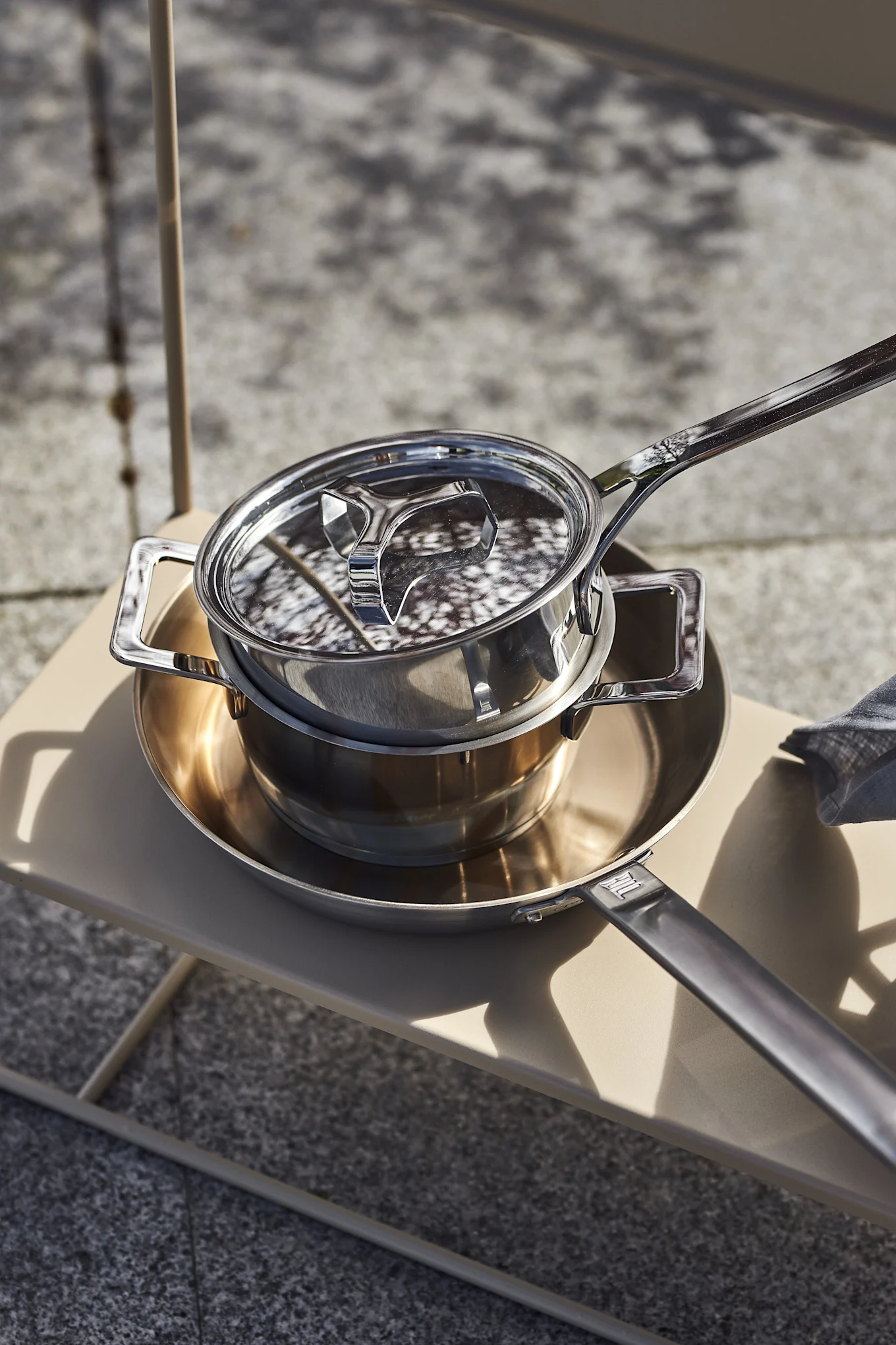 Two stainless steel pots stacked on a beige outdoor table in sunlight.