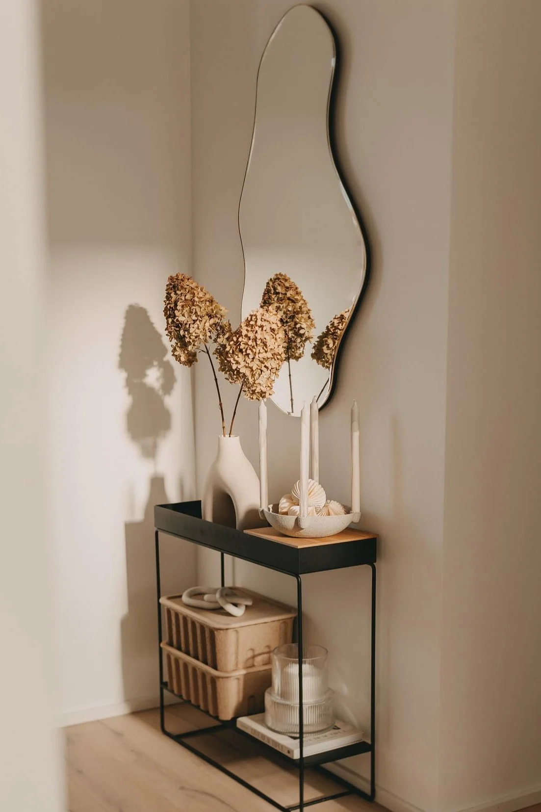 A minimalist console table with a wavy mirror, a vase of dried hydrangeas, candles, and storage boxes.