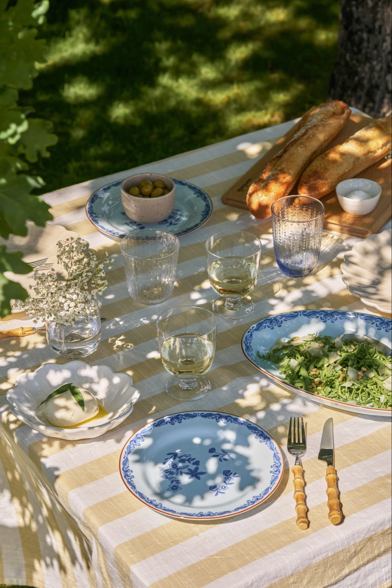 An outdoor picnic table set with a yellow and white striped tablecloth, burrata, salad, baguettes, olives, and blue patterned plates.