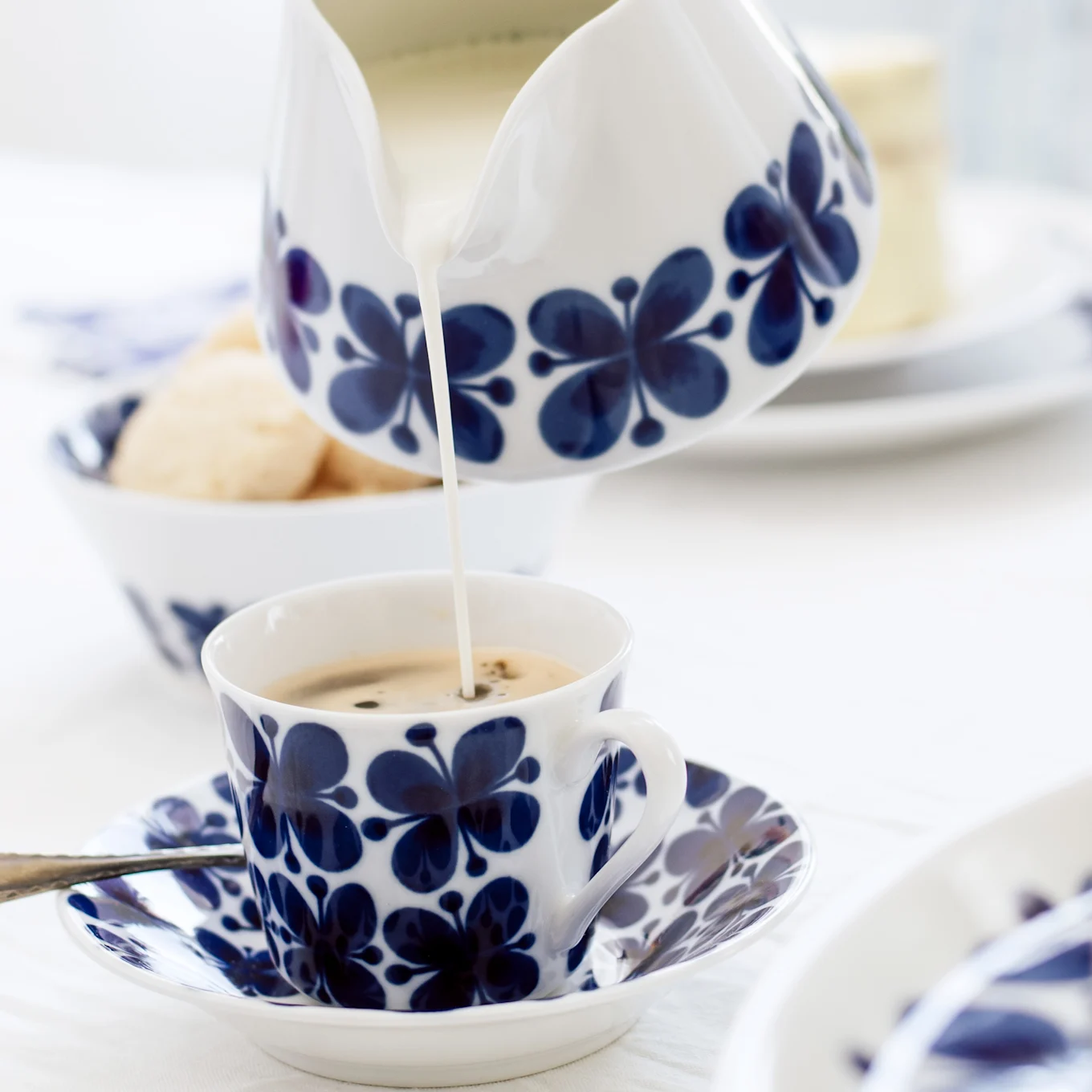 Milk being poured from a blue and white floral creamer into a matching coffee cup on a saucer, with cookies in the background.