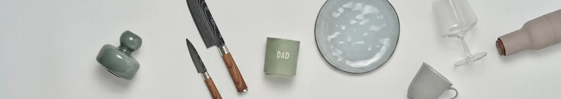 Overhead view of various homeware: a green abstract vase, two knives, a 'DAD' candle, a speckled plate, a wine glass, a grey mug, and a beige bottle on white.