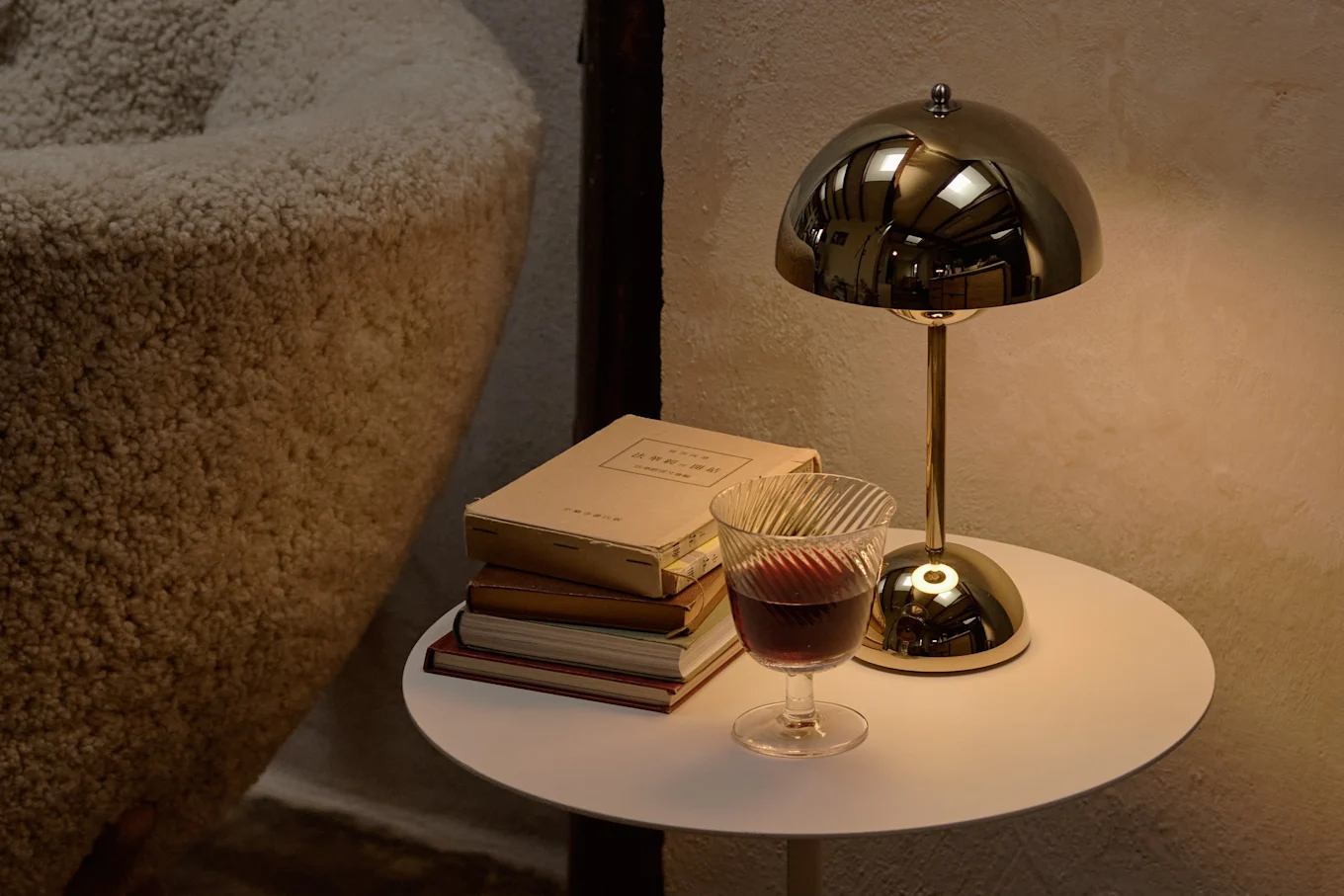 A golden mushroom lamp and stacked books on a white side table with a glass of red wine, next to a textured beige sofa.