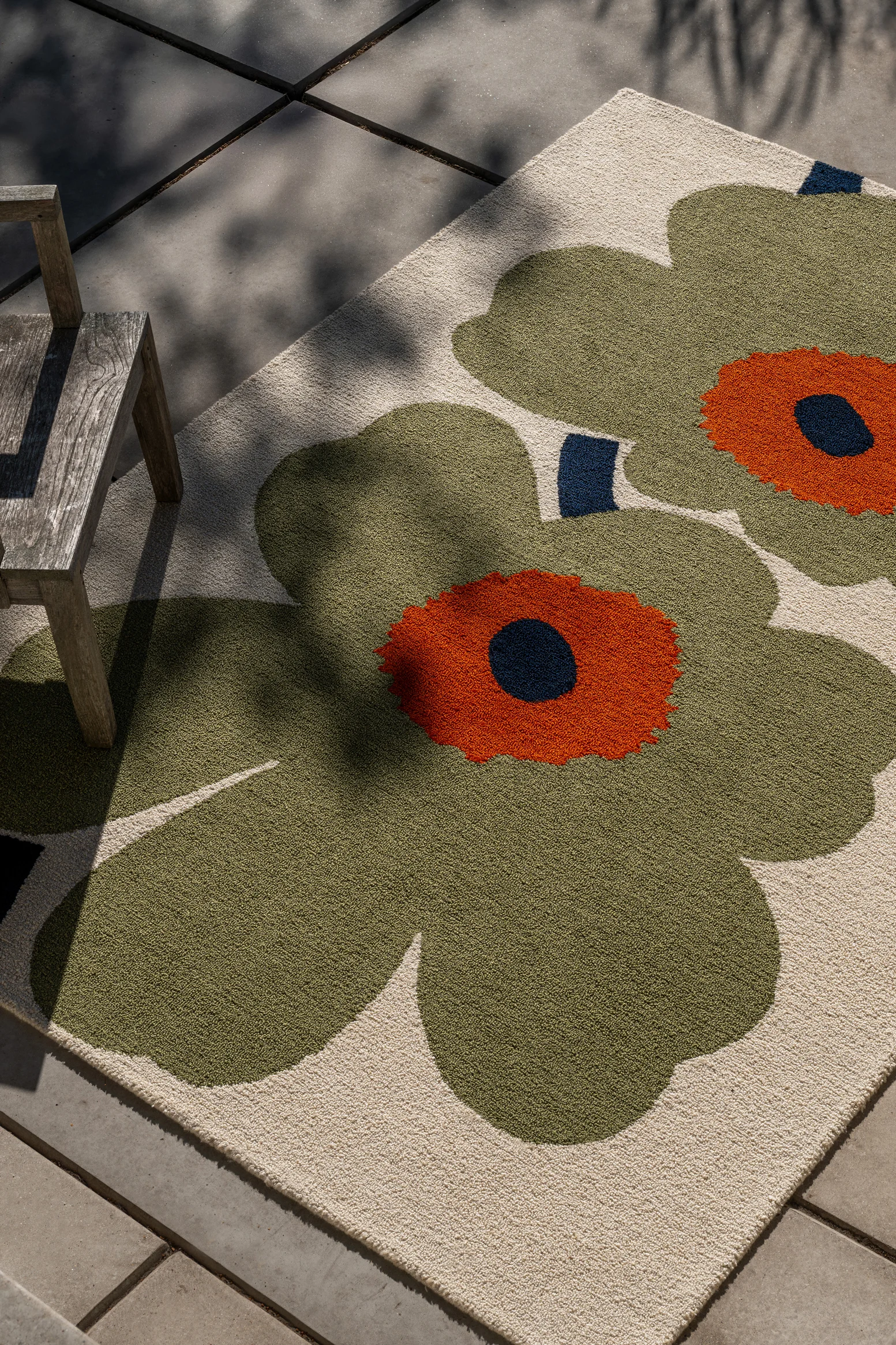 A rug with large green and orange floral patterns on a light background, partially under a wooden chair on a patio, with leaf shadows.