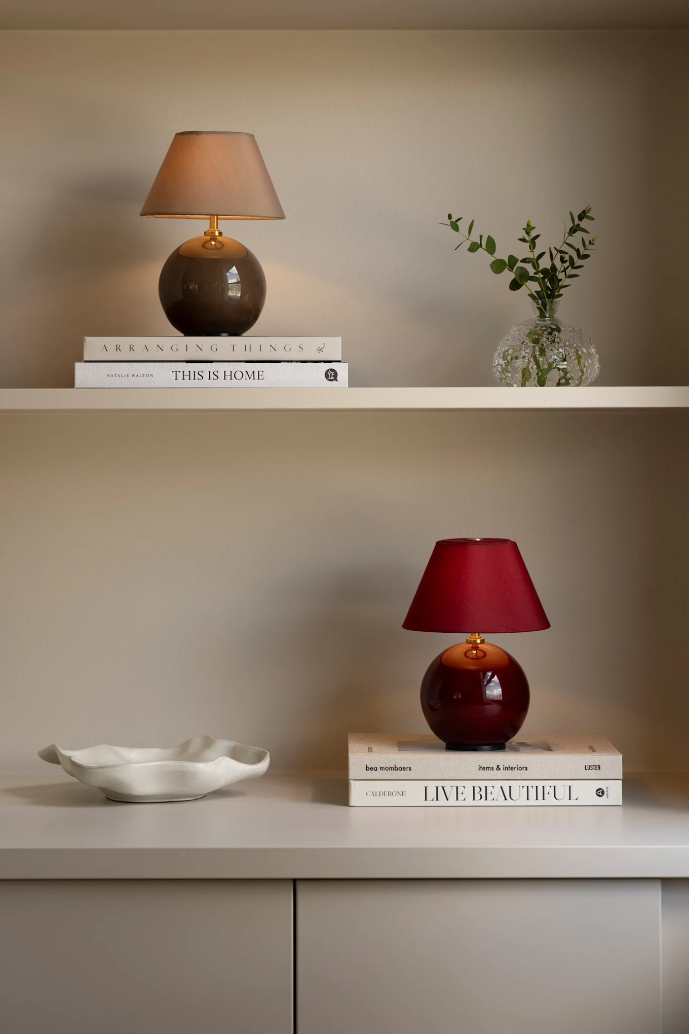 Two shelves displaying decorative items: on top, a beige lamp, books, and a vase with greenery; below, a white bowl, a red lamp, and more books.