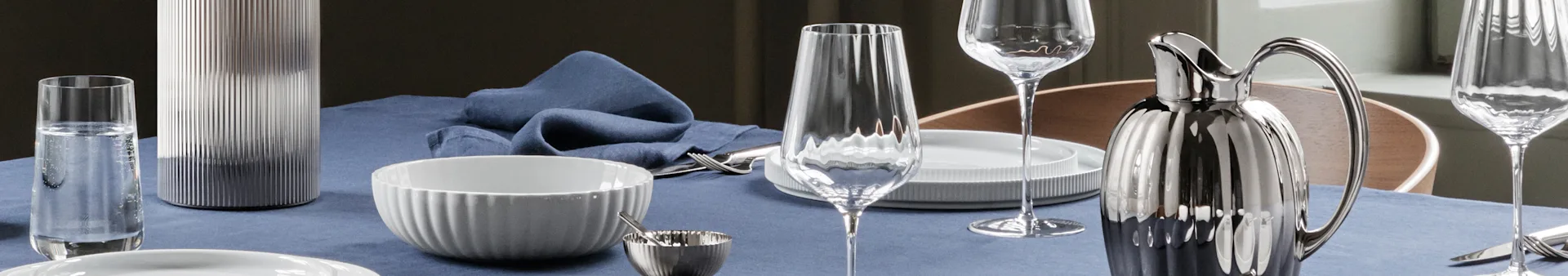 A dining table set with a blue linen tablecloth, featuring white ribbed bowls, clear glasses, silver cutlery, a silver ribbed pitcher, and a grey ribbed vase.