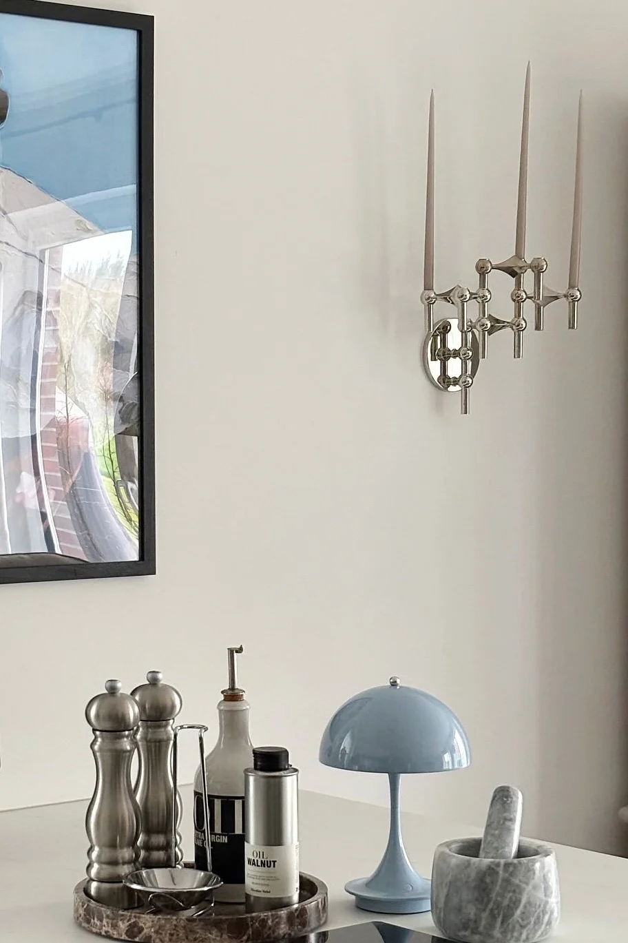 A silver three-candle wall sconce hangs above a white table with kitchen items: grinders, oil bottles, a blue mushroom lamp, and a marble mortar and pestle.