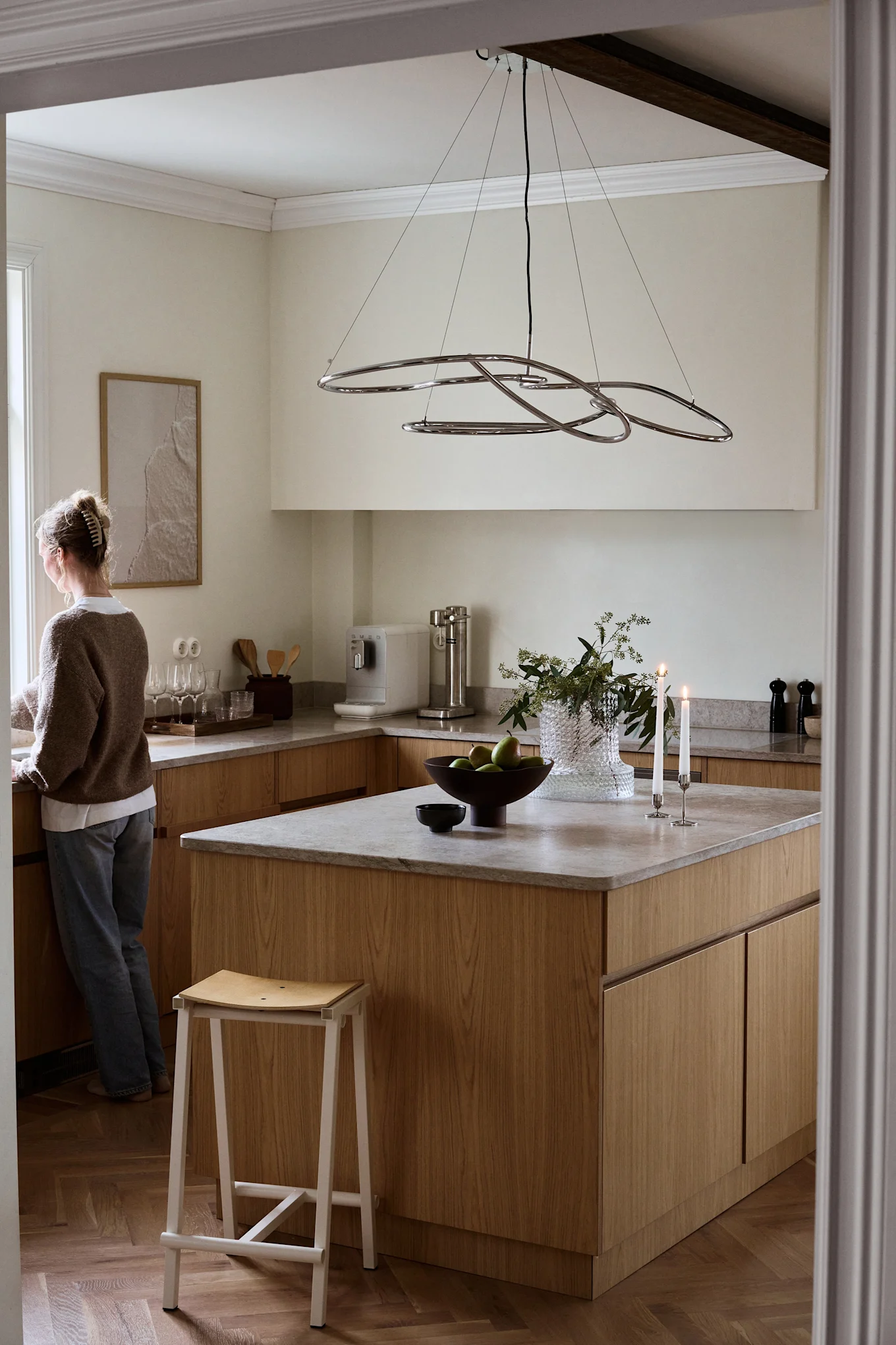A woman stands by a window in a modern kitchen with light wood cabinets, a kitchen island, and a distinctive chrome chandelier.