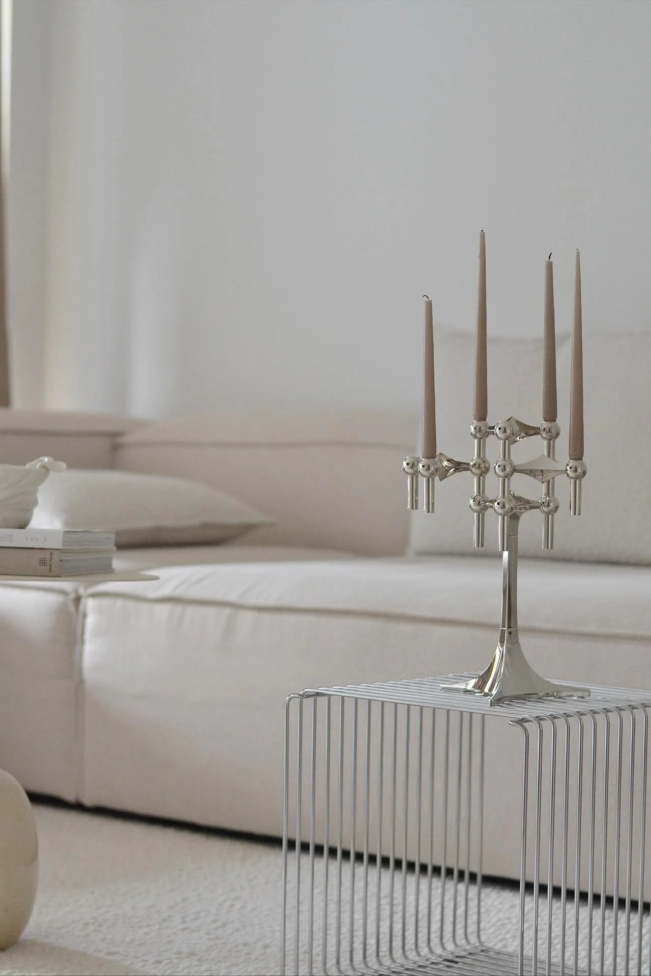 A silver candelabra with five light brown candles sits on a minimalist metal side table. A light-colored sofa is in the background.