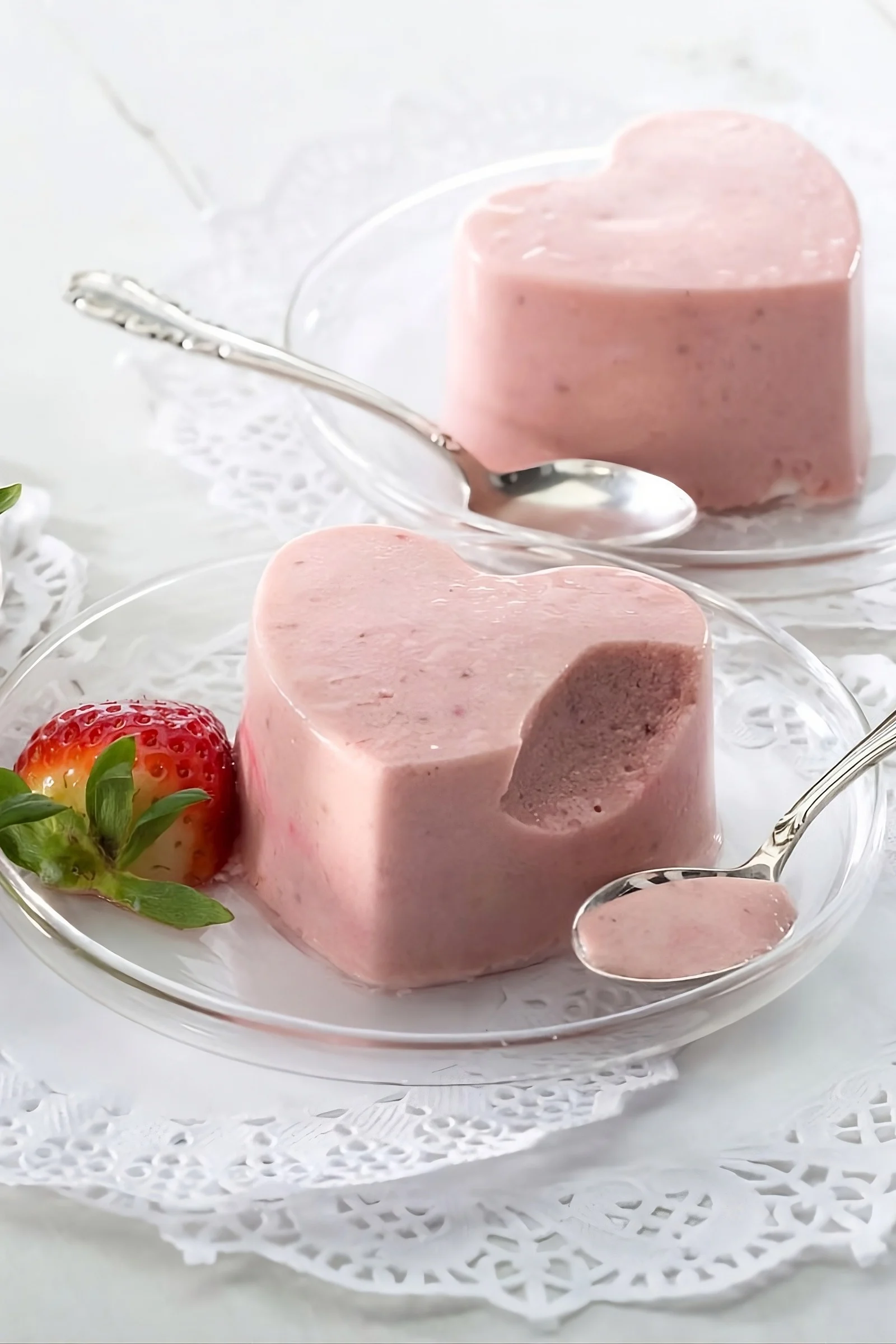Two heart-shaped strawberry desserts on glass plates. One is bitten, revealing soft texture, served with a fresh strawberry and spoons.