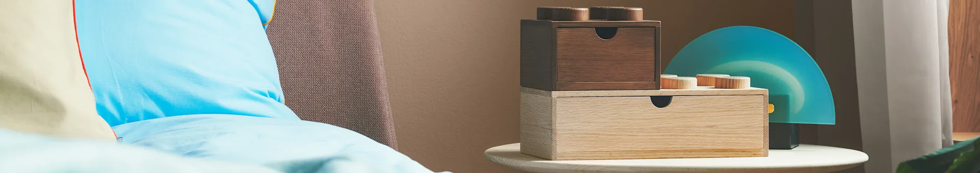 Two stacked, wooden LEGO-style storage boxes with drawers, one dark and one light, on a round table next to a blue decorative panel.