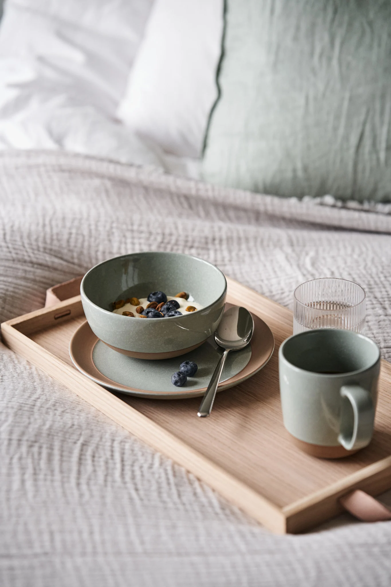 Breakfast in bed: a bowl of yogurt with blueberries and nuts, a spoon, a mug, and a glass of water on a wooden tray.