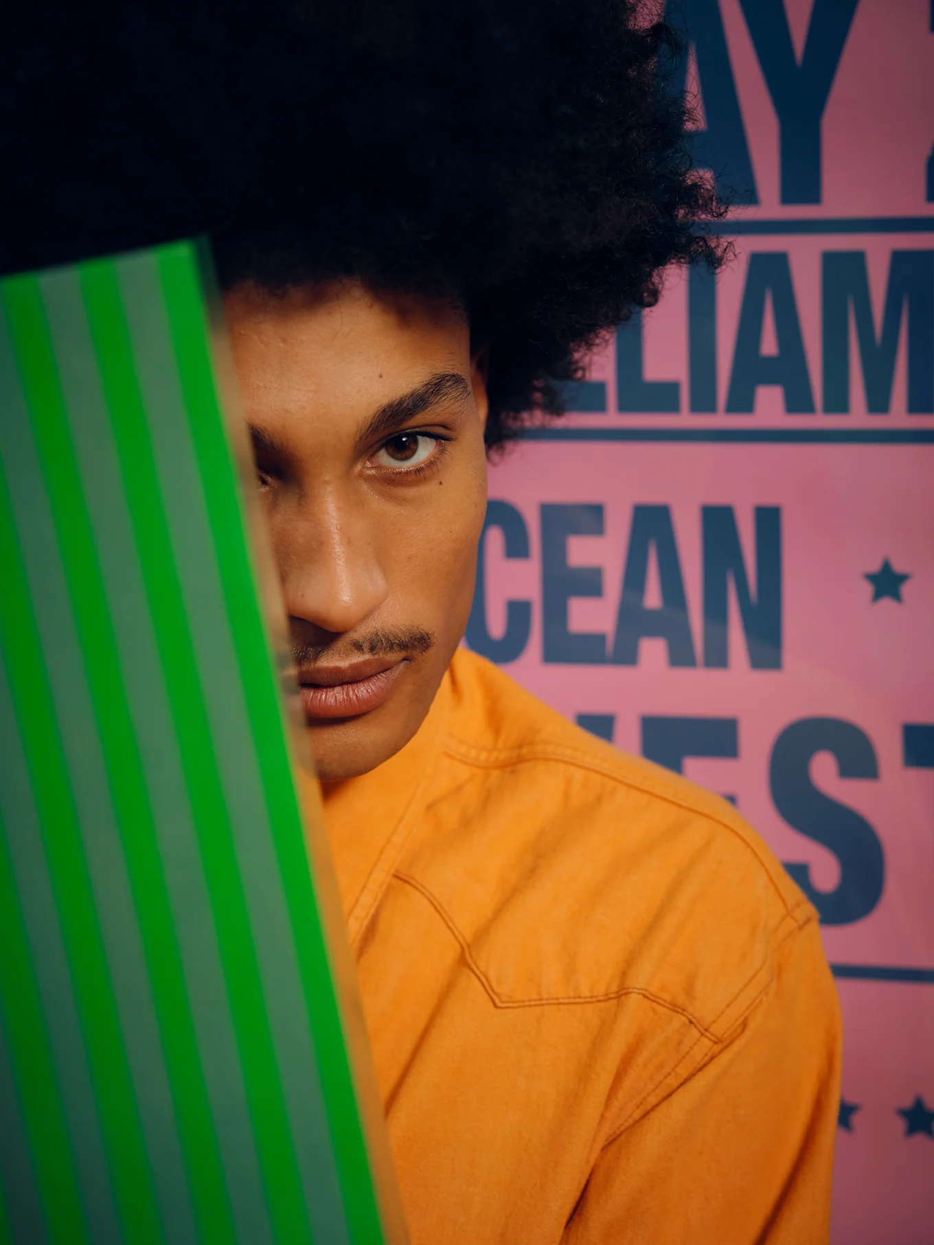 Person with afro, mustache, and orange shirt, peeking from behind a green striped object, looking at camera.