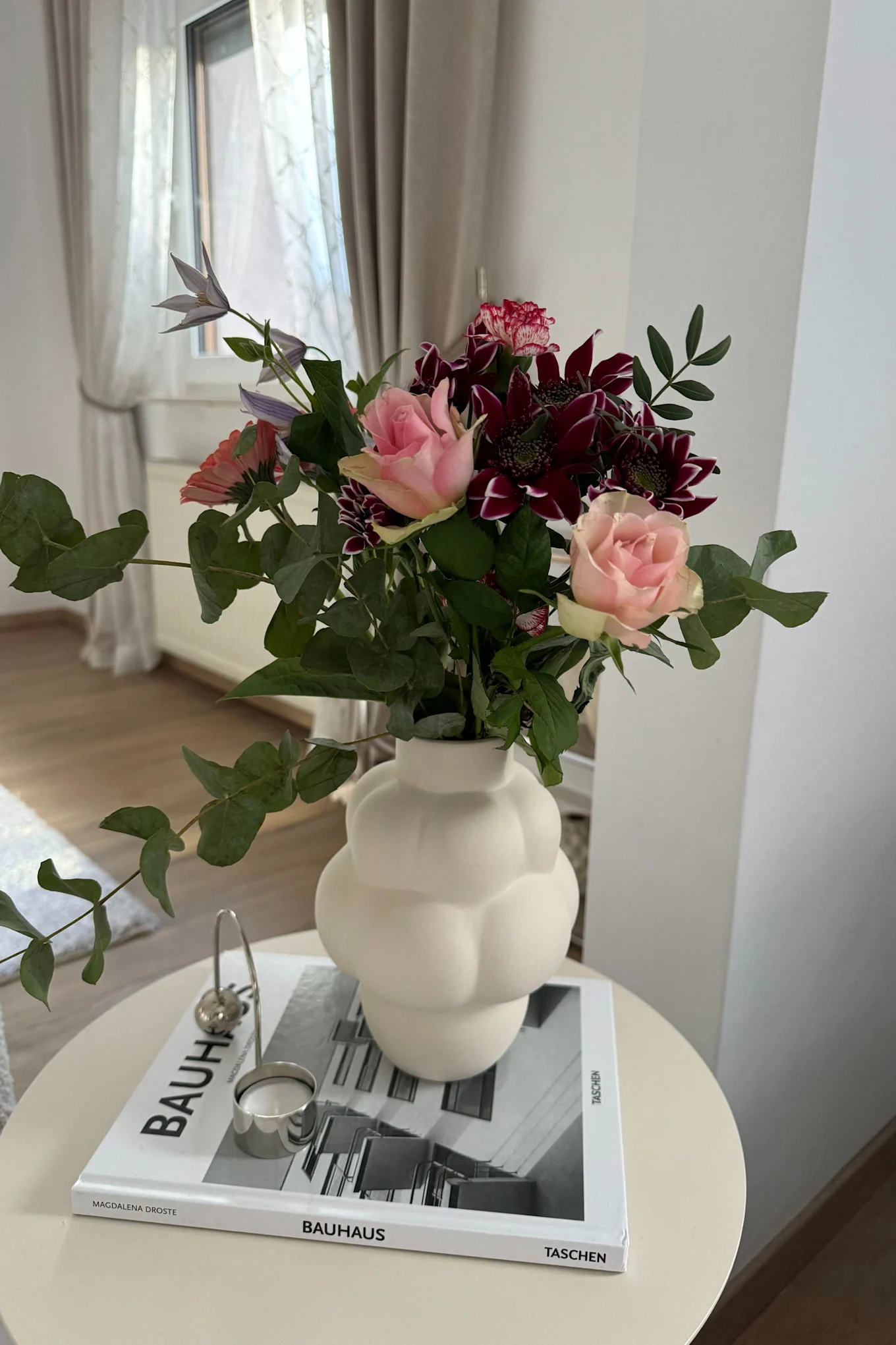 A bouquet of pink roses, dark red flowers, and eucalyptus in a white sculptural vase on a Bauhaus book.