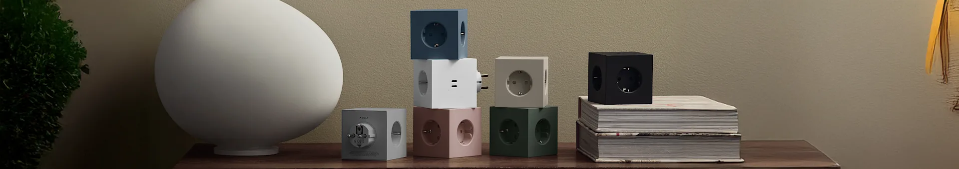 Colorful cube-shaped power strips in blue, white, grey, pink, green, and black on a wooden table next to a white lamp and books.