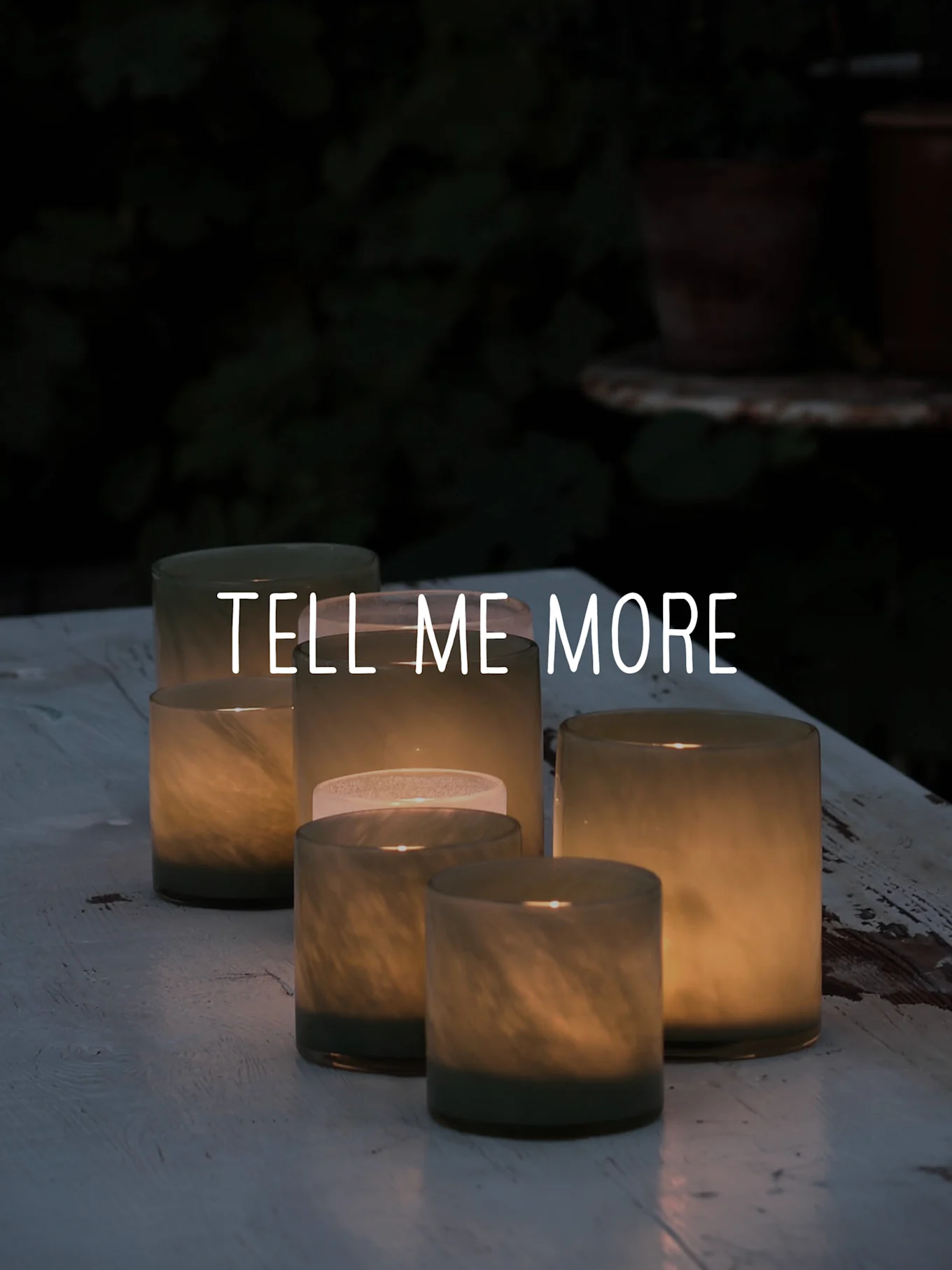 Frosted glass candle holders with lit candles on a rustic white table outdoors, overlaid with 'TELL ME MORE'.