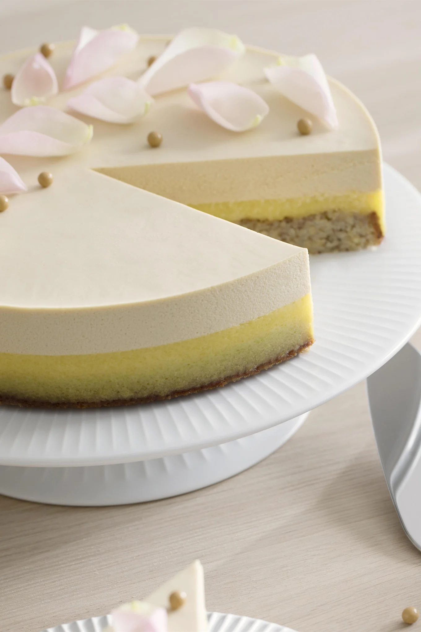 Multi-layered cake with a slice removed, decorated with pink petals and golden balls, on a white cake stand.