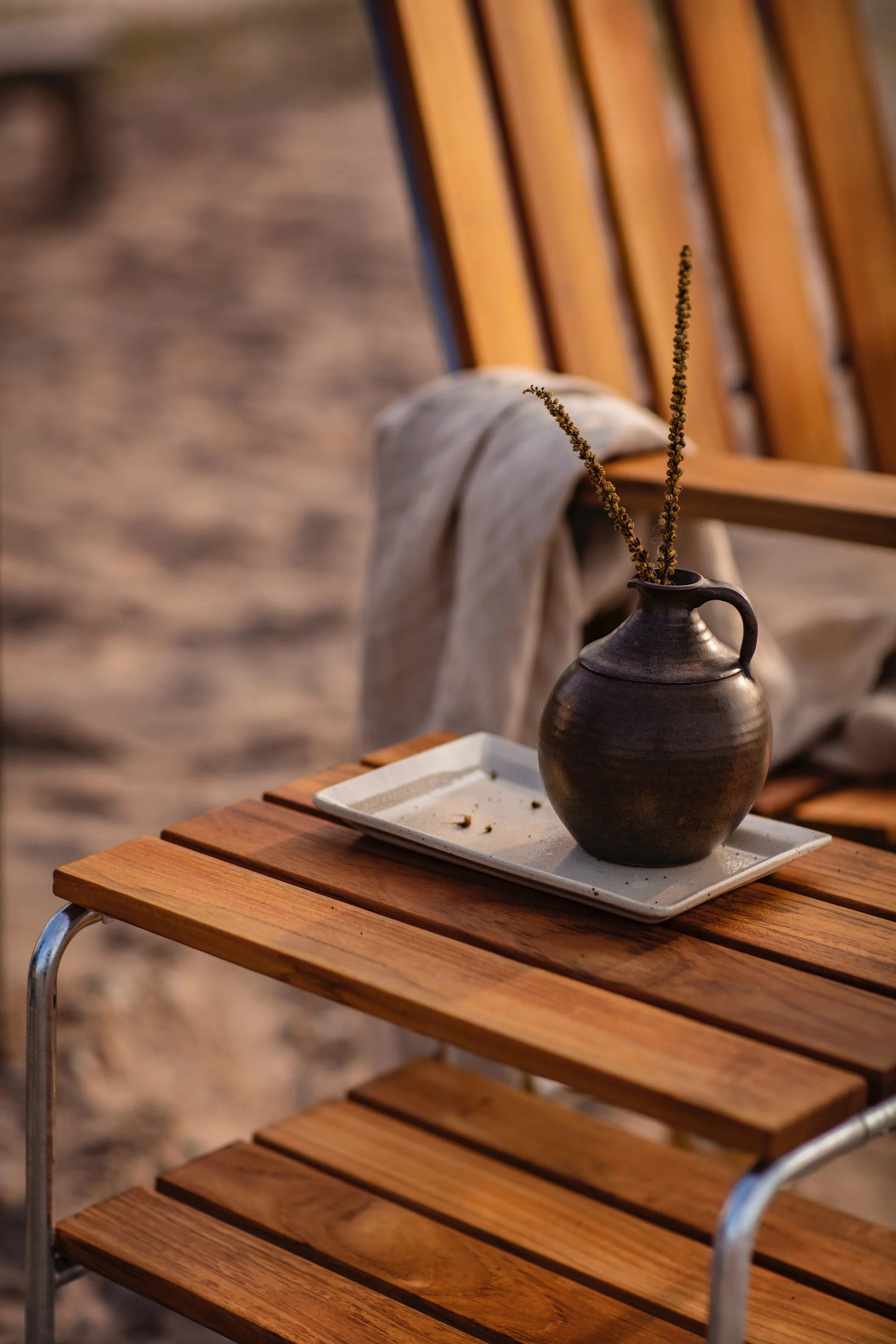 Dark ceramic vase with dried stems on a white tray atop a wooden slatted table outdoors.