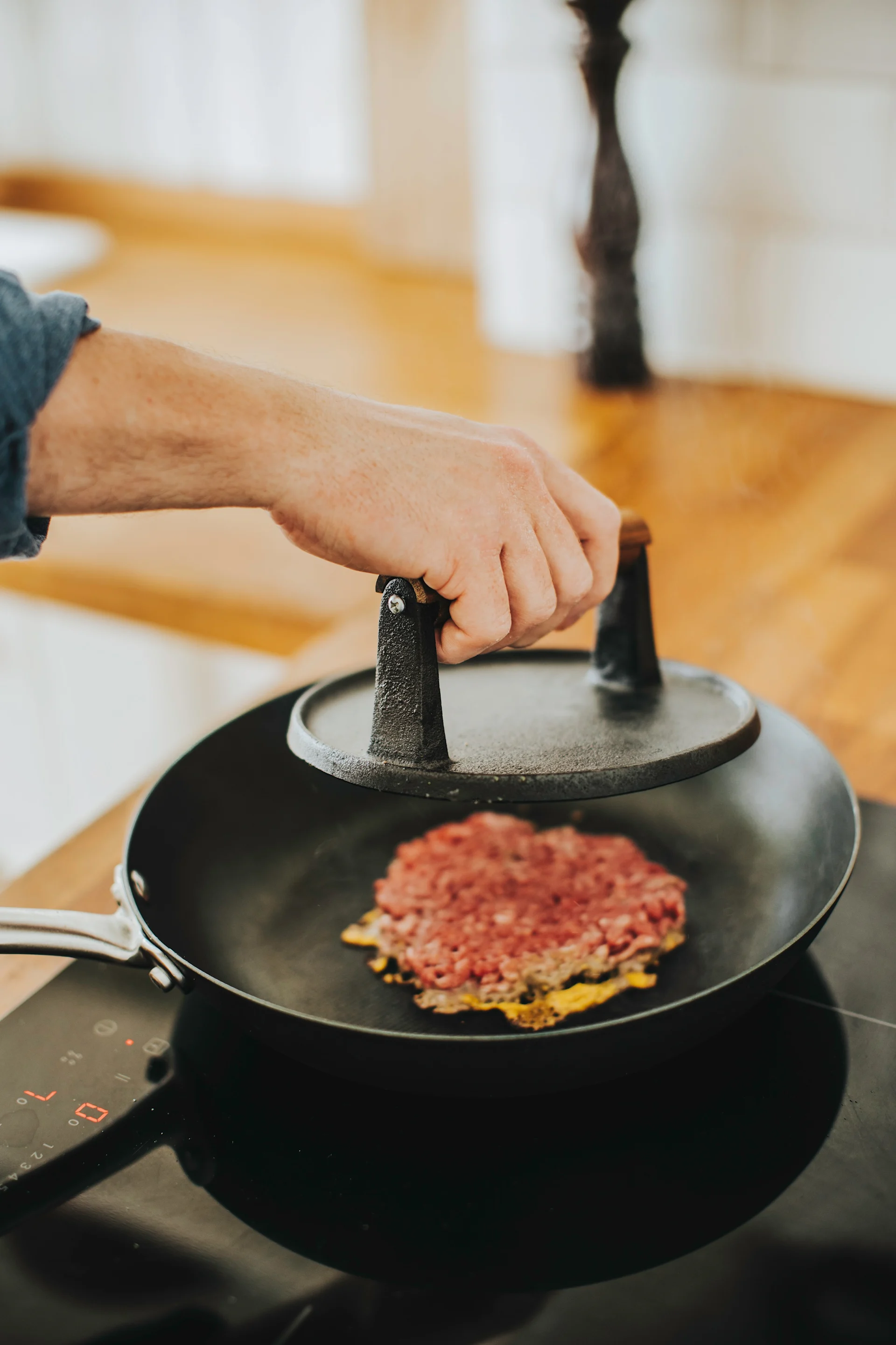 Hand pressing a raw burger patty in a frying pan with a burger press on an induction stovetop.