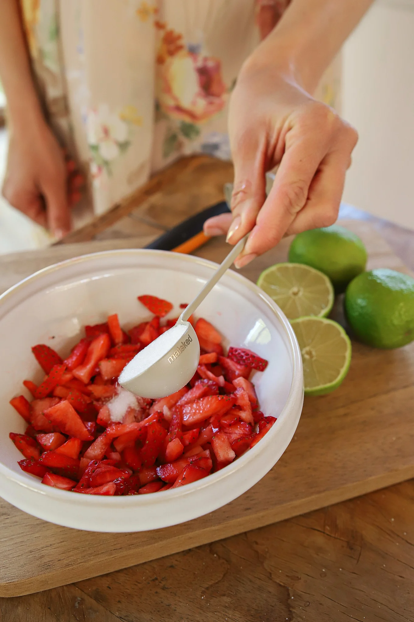 Hand adding sugar with a measuring spoon to chopped strawberries in a white bowl, with limes on a wooden board.