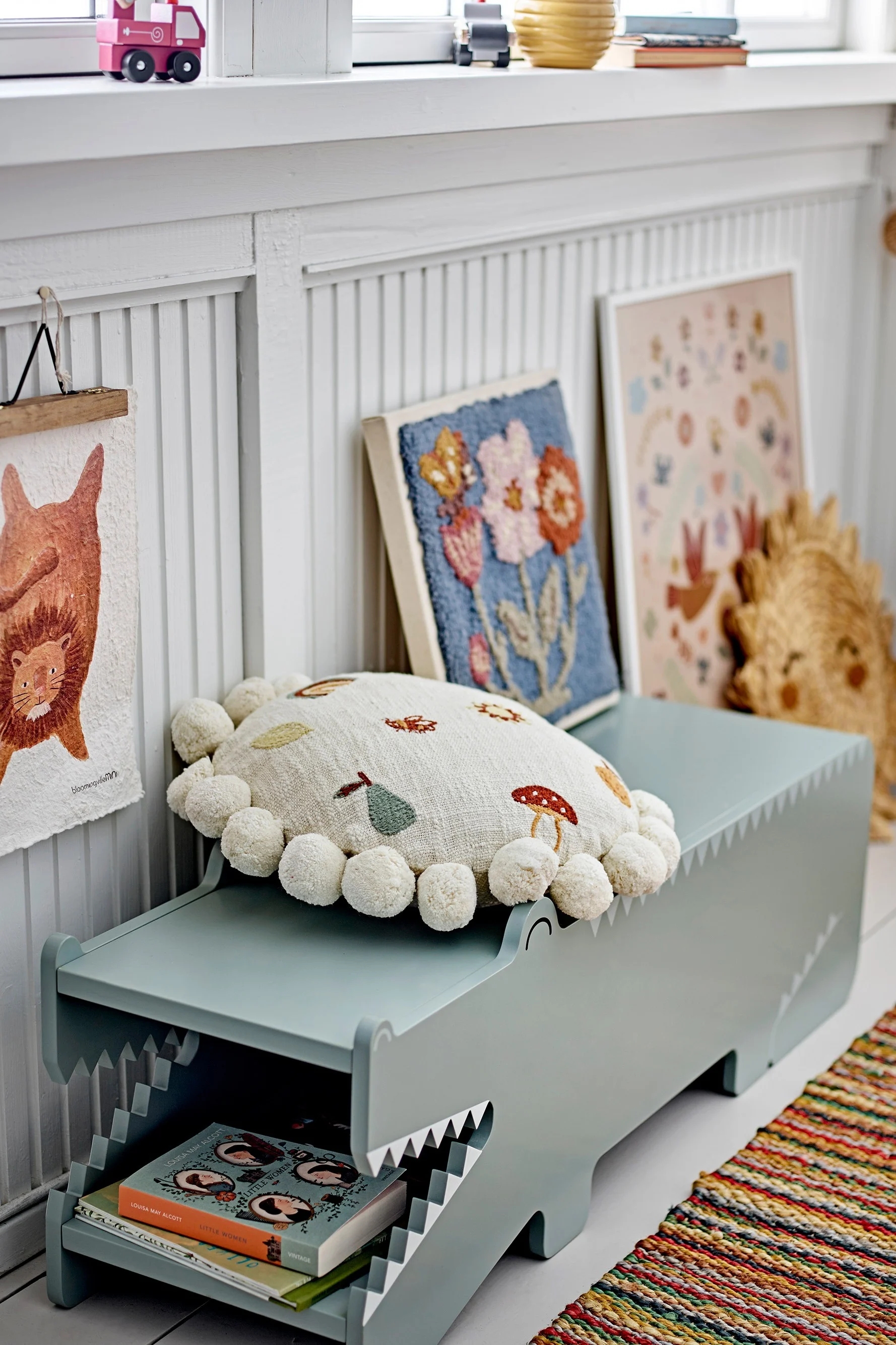 Light green crocodile-shaped storage bench with a pom-pom cushion and books in its mouth. Artwork hangs on a white paneled wall.