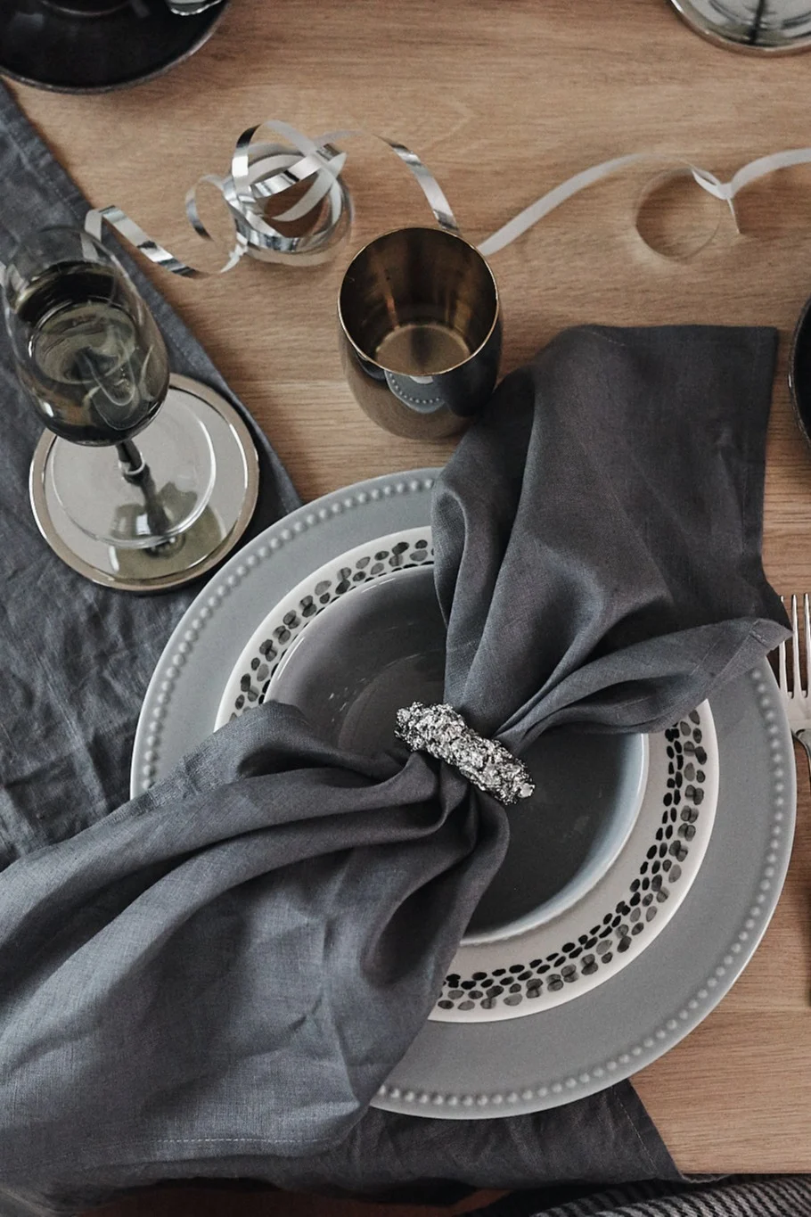 Elegant grey table setting with stacked plates, linen napkin in a sparkly ring, wine glass, and silver streamers.