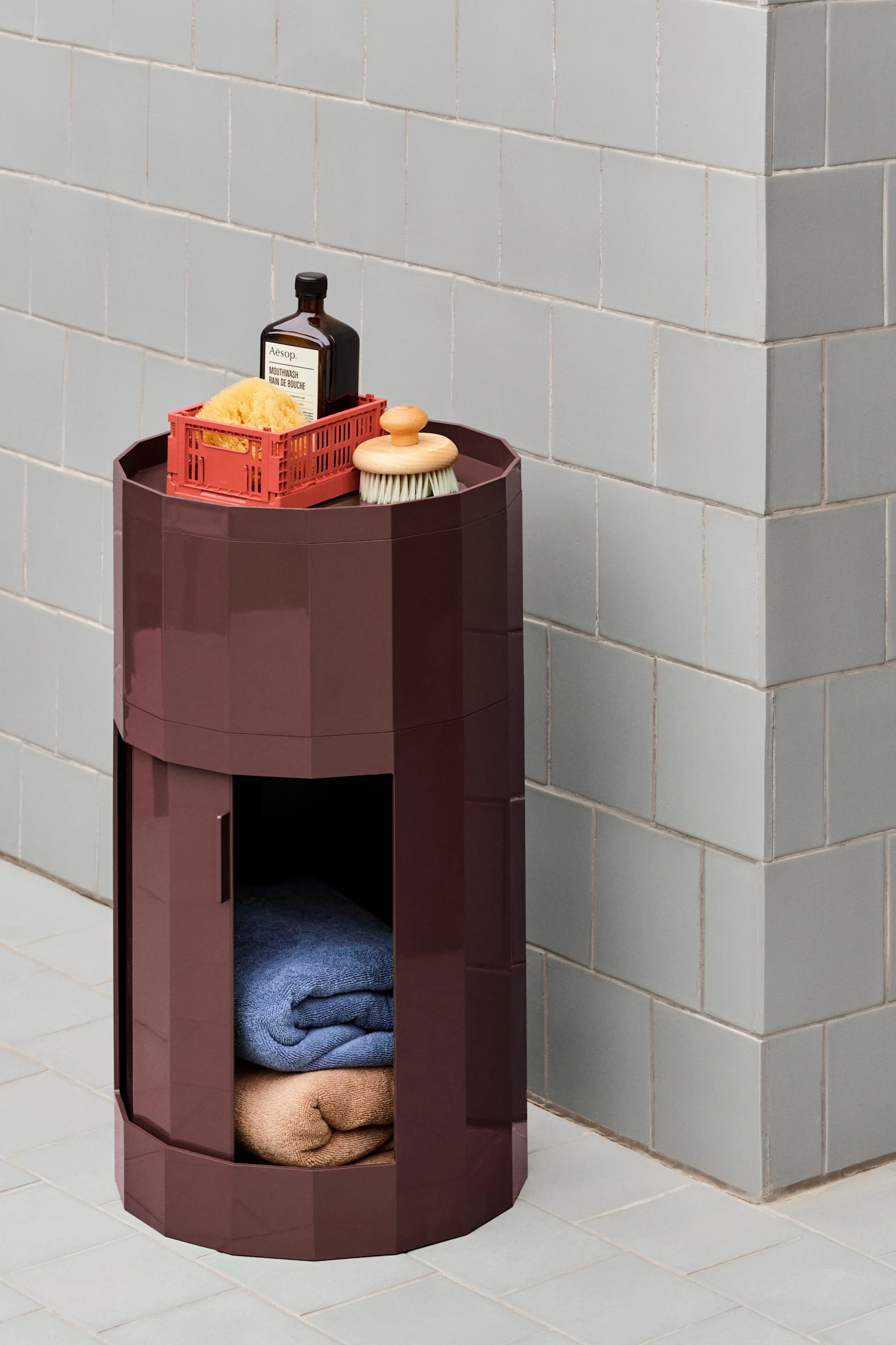 A dark red hexagonal storage unit with bath products on top and folded towels inside, against a light grey tiled wall.