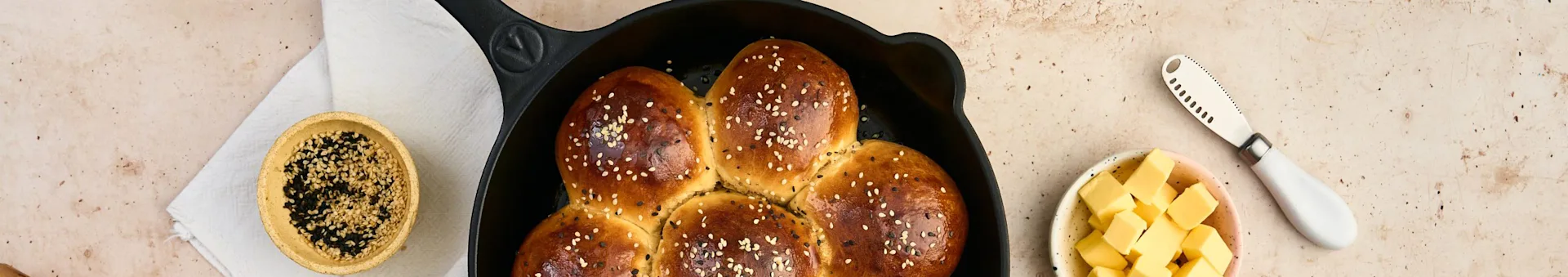 Golden-brown bread rolls with sesame seeds in a black skillet, next to a bowl of sesame seeds, cubed butter, and a knife.