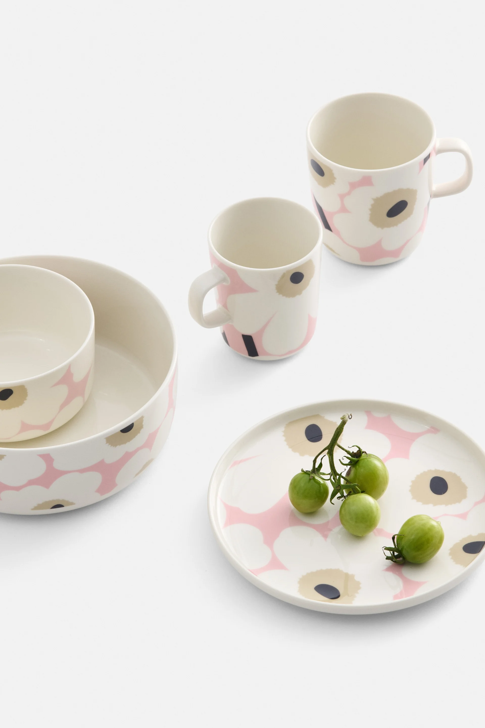 Cream-colored dinnerware with pink and black floral pattern, including two bowls, two mugs, and a plate with green cherry tomatoes.